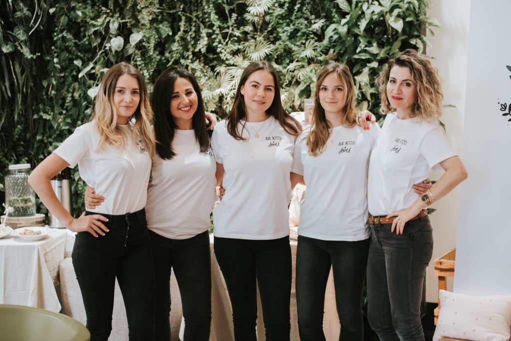 A group of five women standing together, wearing matching white t-shirts and black jeans, in front of a lush green wall with plants.